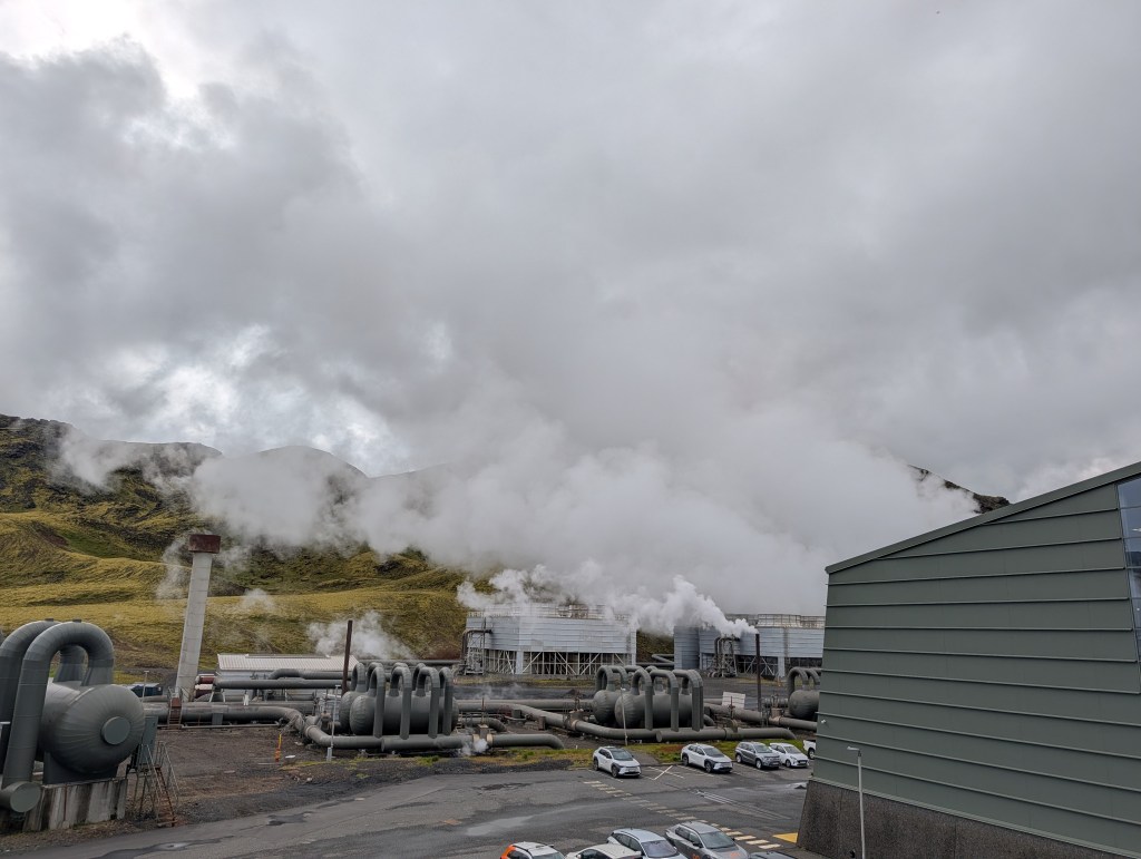 Industrial geothermal power plant with steam rising against a cloudy sky and hilly landscape.