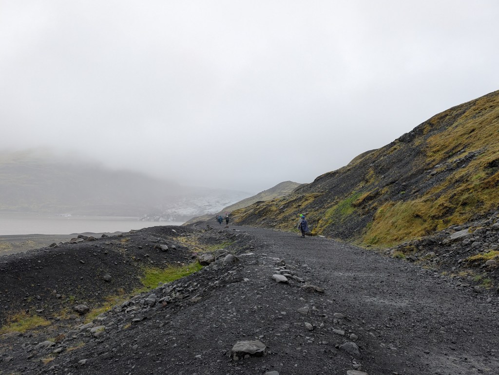 Two hikers walking along a rocky trail in a foggy landscape with mountains and a glacier in the background.