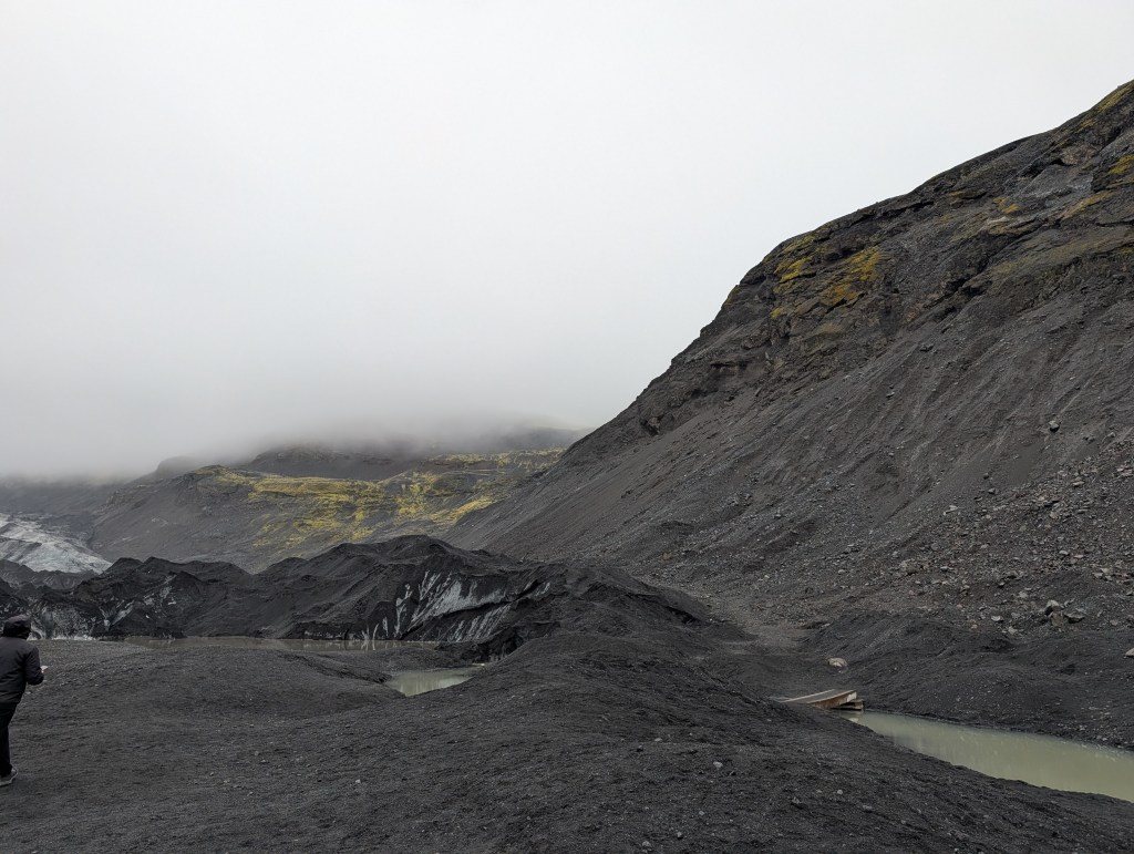 A rugged landscape featuring dark volcanic rock and mist, with a person standing on a path near a small body of water.