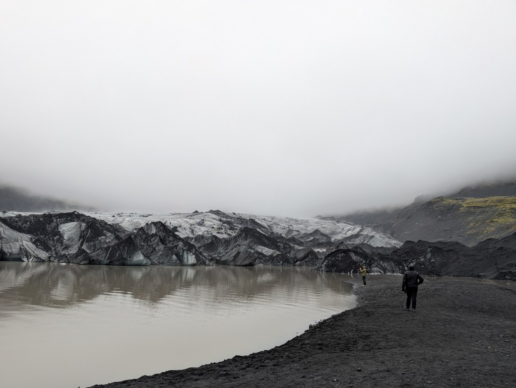 A scenic view of a glacier at a lake surrounded by rocky terrain and misty mountains, with two people standing on the shore.