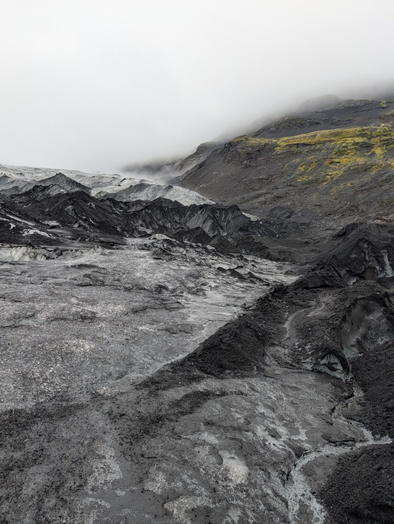 A rugged landscape featuring dark volcanic rock and glacial features, partially obscured by low clouds and mist.