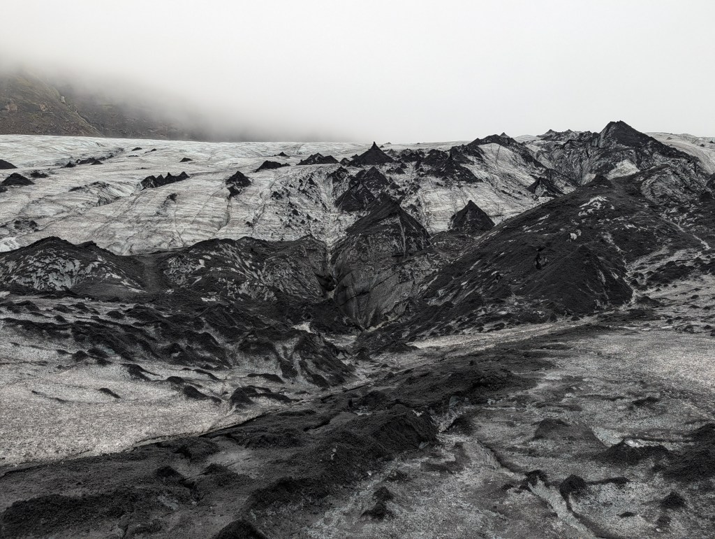 A rocky glacier landscape with dark volcanic rock and icy surfaces under a cloudy sky.