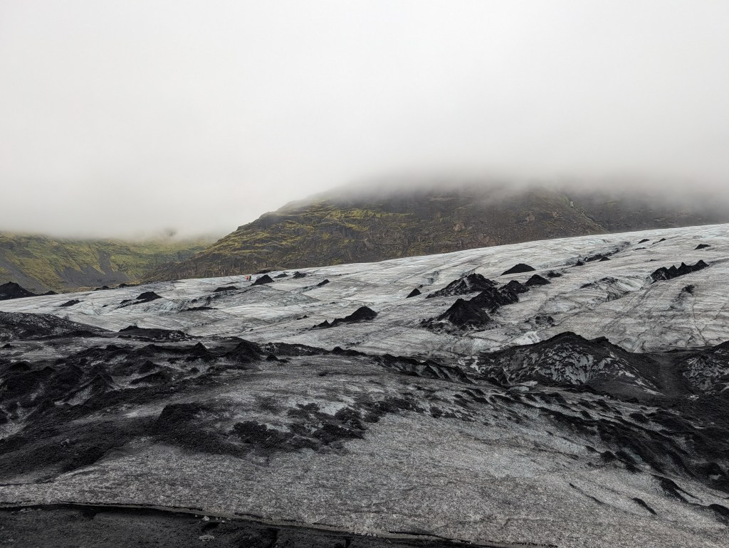 A landscape of a glacier with textured ice and black volcanic rock formations, surrounded by misty mountains and overcast skies.