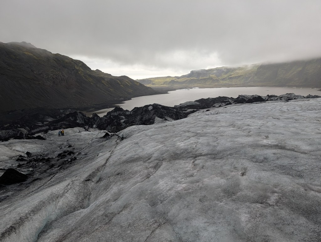 A panoramic view from a glacier overlooking a dark, rocky terrain leading to a calm lake, with two small figures in the distance.