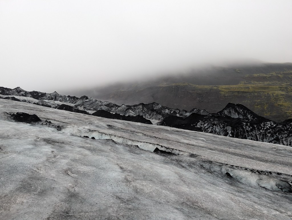 A view of a glacier's icy surface with jagged black and gray textures, under a foggy sky, revealing a mountainous landscape in the background.