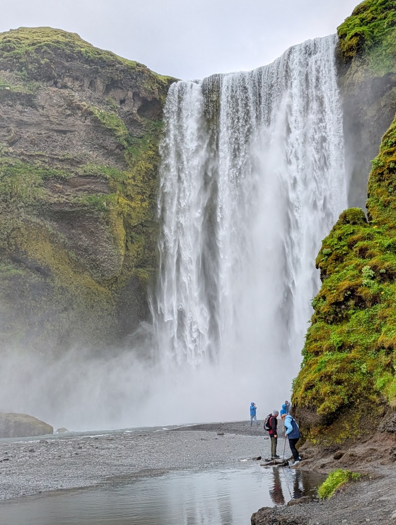 A majestic waterfall cascading down a rocky cliff surrounded by lush greenery, with several people standing on a rocky beach in the foreground.