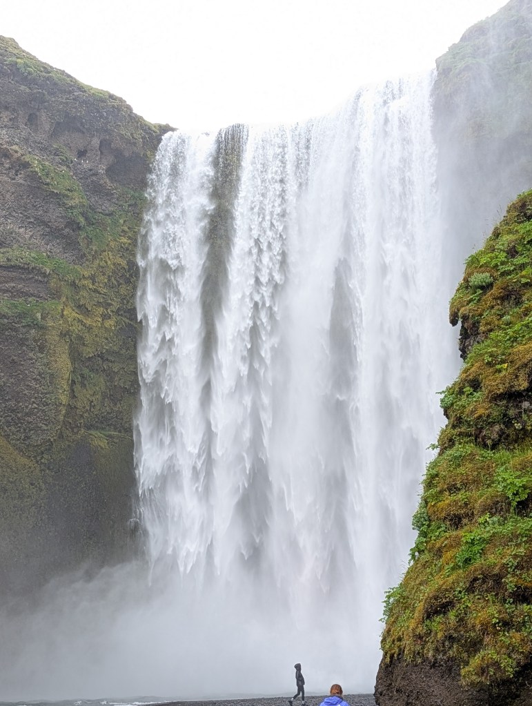 A majestic waterfall cascading down a rocky cliff surrounded by lush greenery, with two people standing near the water's edge.