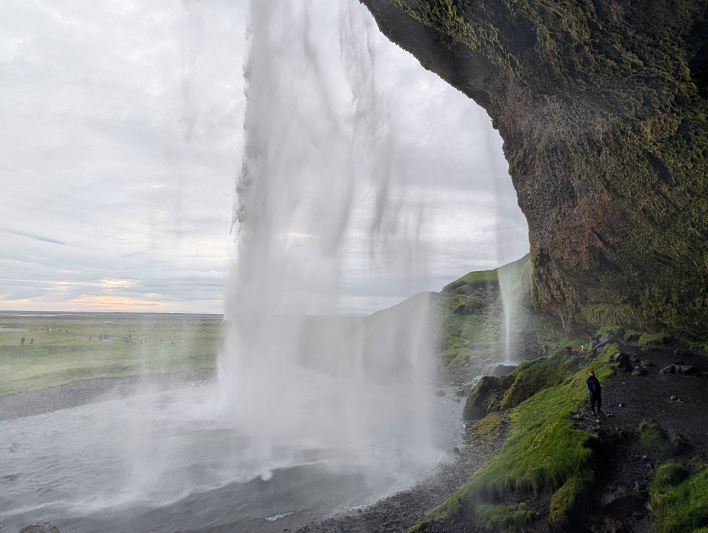 A person stands near the base of a waterfall, with water cascading over a rocky ledge. The scene is set in a grassy landscape under a cloudy sky, showcasing the natural beauty of the area.