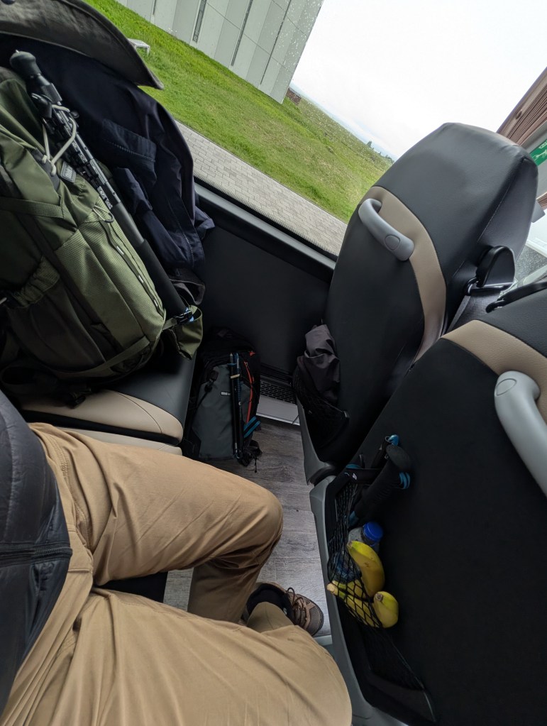 Interior of a vehicle showing two pairs of pants, a collection of backpacks, and items in the seat pocket, including a bottle and bananas.