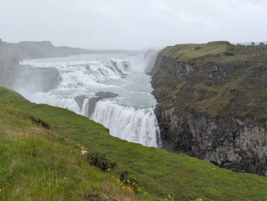 A panoramic view of a majestic waterfall cascading over rocky cliffs, surrounded by lush green grass and mist, under a cloudy sky.