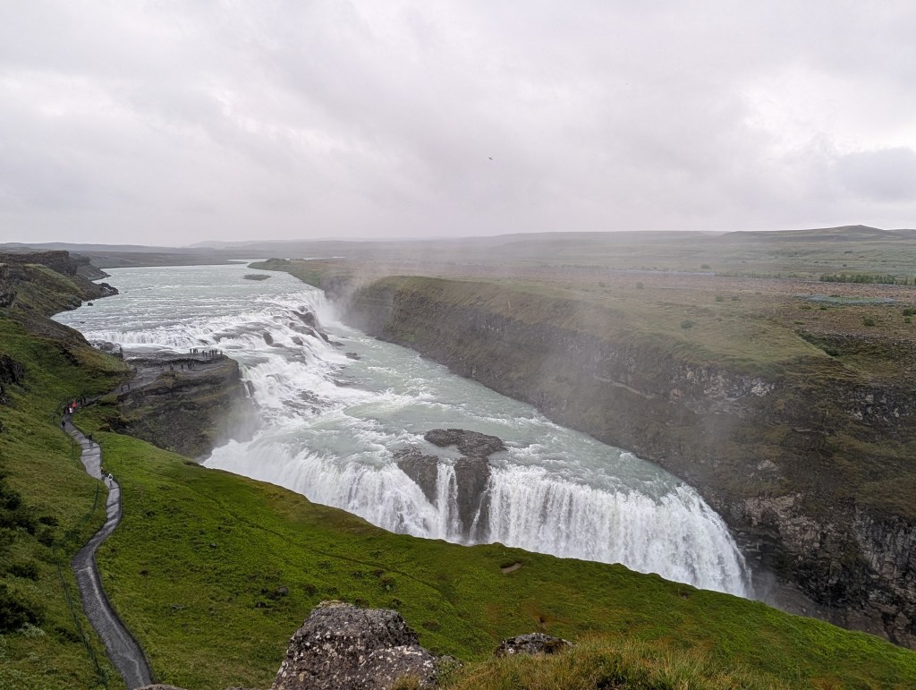 A panoramic view of a powerful waterfall cascading into a river, surrounded by green hills and a cloudy sky.