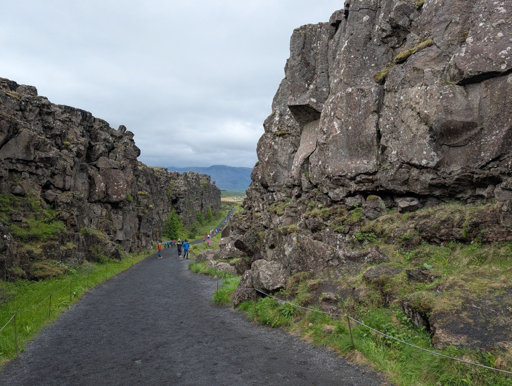 A narrow path between towering volcanic rock formations, with a group of hikers in colorful clothing walking along the trail under a cloudy sky.