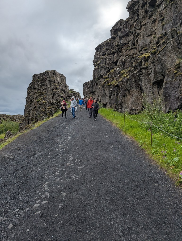 A walkway surrounded by towering rock formations with several people walking along it on a cloudy day.