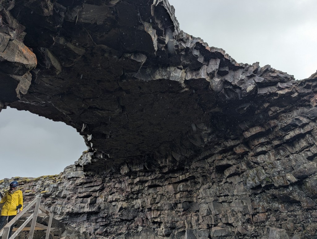 A person in a yellow jacket stands near a large, rocky arch formation under a cloudy sky.