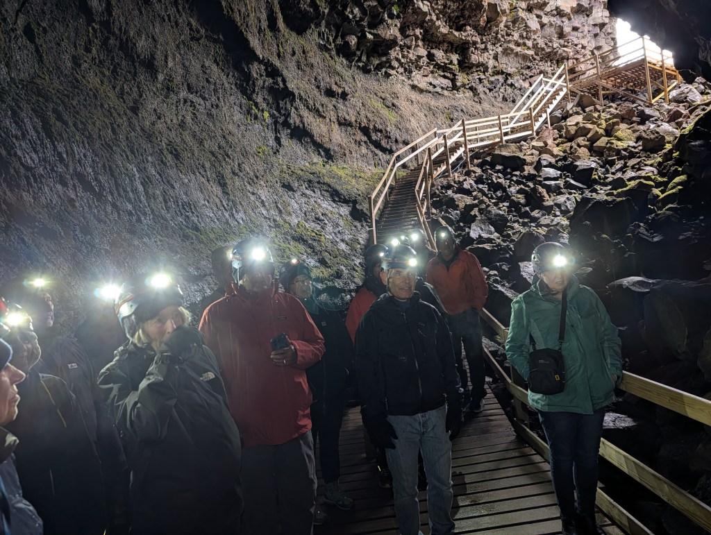 A group of people wearing headlamps stands inside a cave, with a rocky texture on the walls and a wooden walkway leading into the distance.