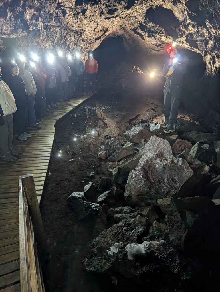 A guided group tour inside a dark cave, with participants wearing headlamps and standing on a wooden path, while a guide stands near a pile of rocks.