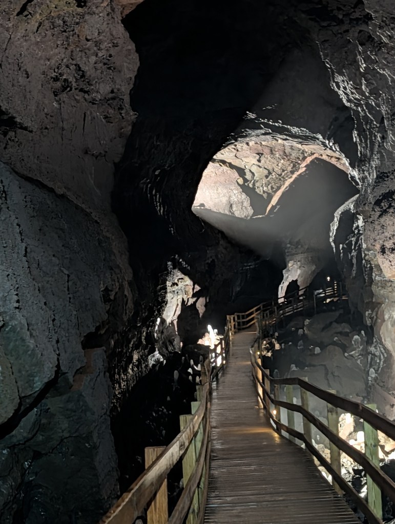 A dimly lit cave interior featuring a wooden walkway leading through rocky formations, with beams of light illuminating the space.