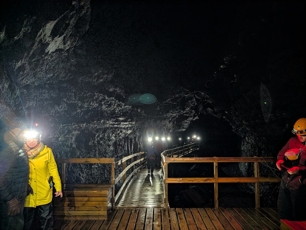 Interior of a dark cave with a wooden walkway, illuminated by headlamps worn by visitors.