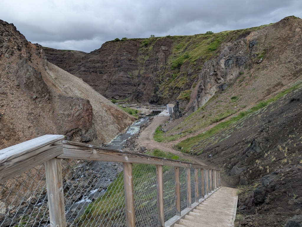 A wooden staircase leading down into a canyon with rocky and grassy slopes, flanked by a small stream running through the valley.