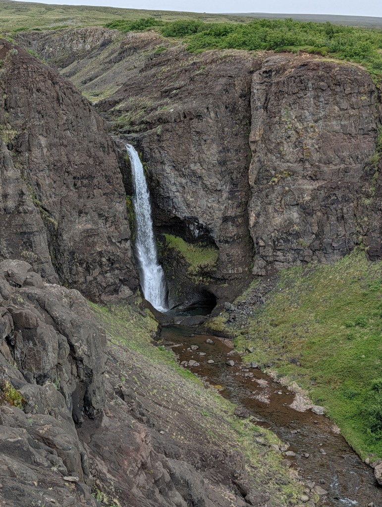 A waterfall cascading down rocky cliffs into a small river surrounded by lush greenery.