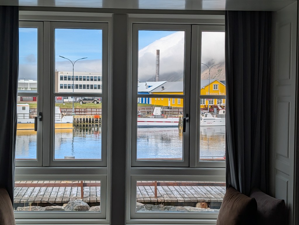View of a colorful harbor scene through a large window, featuring boats and buildings, with a backdrop of mountains and blue sky.