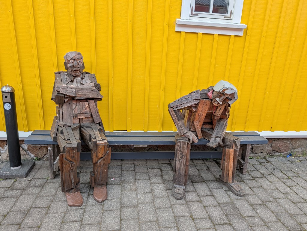 Two wooden sculptures of elderly men sitting on a bench in front of a bright yellow wall.