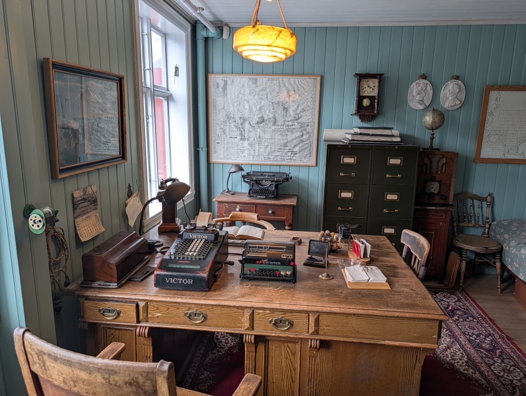 A vintage office interior featuring a wooden desk with a typewriter and various stationery items. The room has light blue walls, a globe, framed maps, and an antique clock, creating a nostalgic atmosphere.