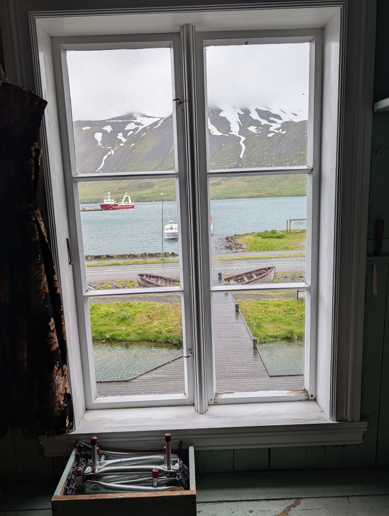 View from a window showing a scenic landscape with mountains, a body of water, and boats. The window frame is white, and there is a box with metallic objects in the foreground.