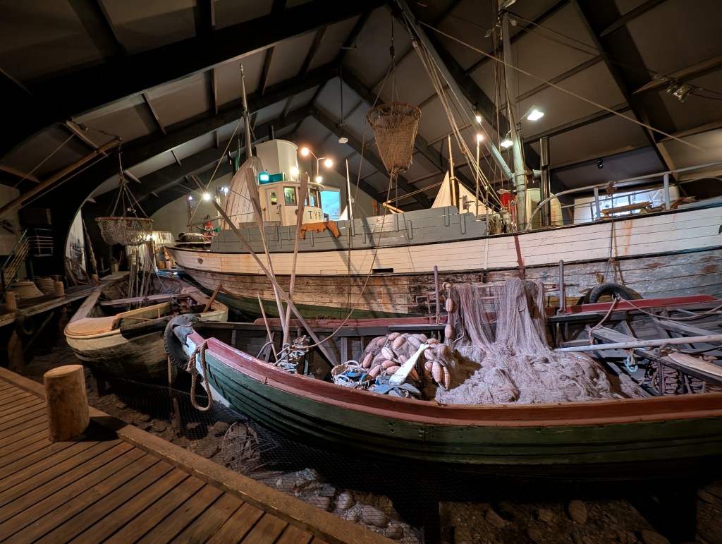 Multiple vintage fishing boats are displayed inside a large indoor exhibit, with nets and fishing equipment present, highlighting maritime heritage.