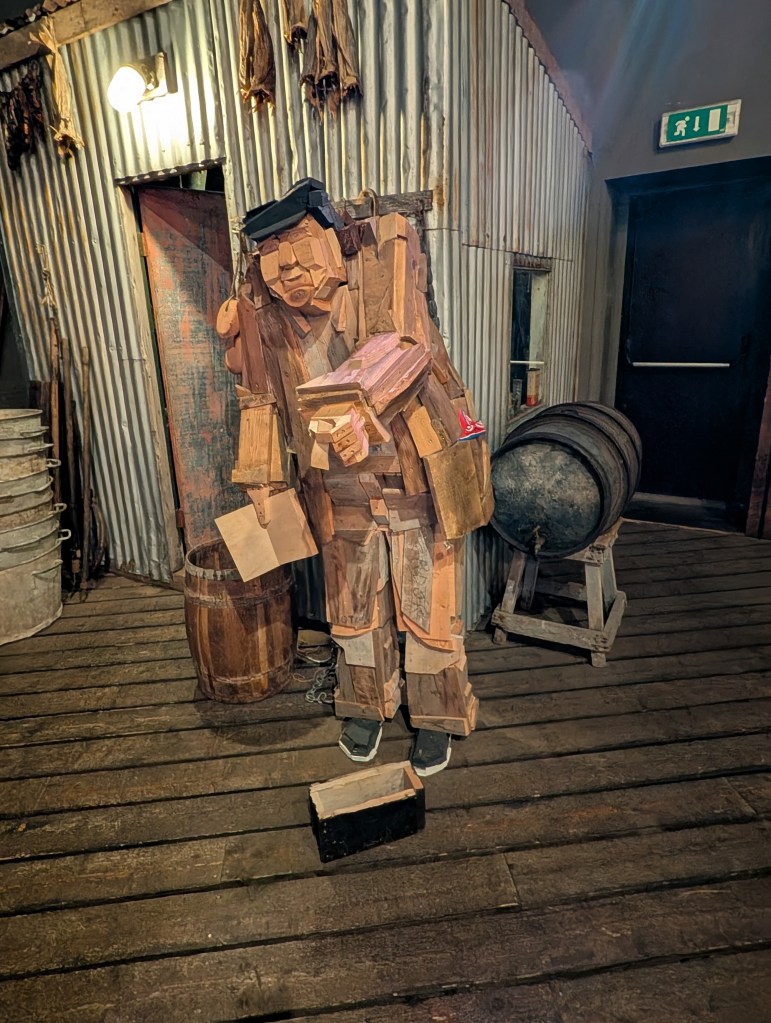 A wooden sculpture depicting a person made of various pieces of wood, standing in an industrial setting with a corrugated metal wall and wooden barrels in the background.