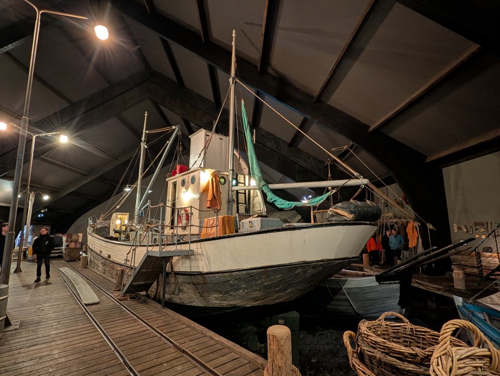 An old fishing boat displayed inside a large, well-lit exhibit hall, with wooden flooring and various items surrounding it, including baskets and smaller boats.