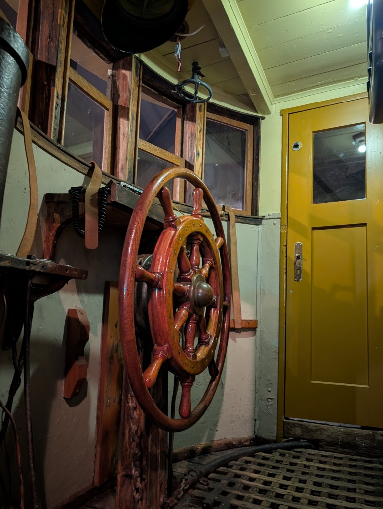 The interior of a boat featuring a wooden ship's wheel, with a yellow door in the background and wooden walls surrounding it.