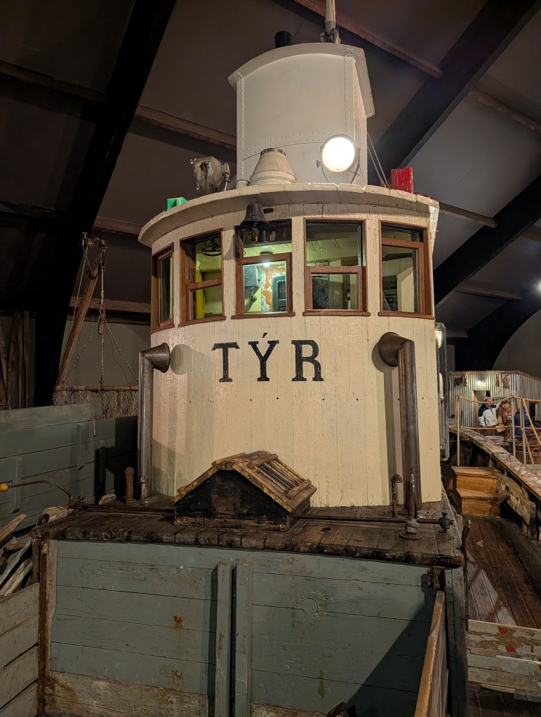 A close-up view of the vintage tugboat 'TÝR' displayed indoors, highlighting its distinct cabin with windows and a light on top.