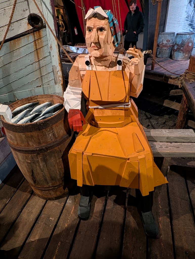 A large wooden sculpture of a woman wearing an orange dress and gloves, sitting on a wooden platform beside a barrel filled with fish.