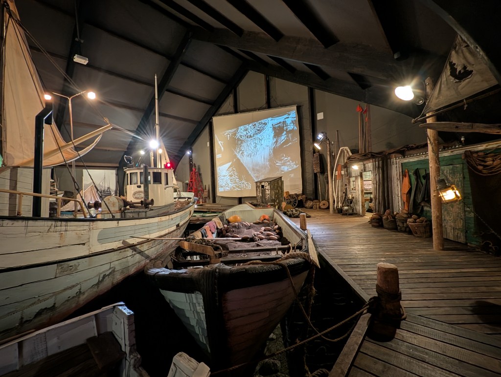 Interior of a maritime museum featuring vintage boats, a large projection screen, and various fishing equipment displayed in a rustic setting.