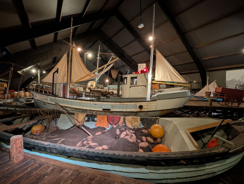 A view of a marine museum featuring historic boats, including a large sailing vessel with a tan sail and a smaller boat with fishing gear and nets, all situated under a high ceiling.