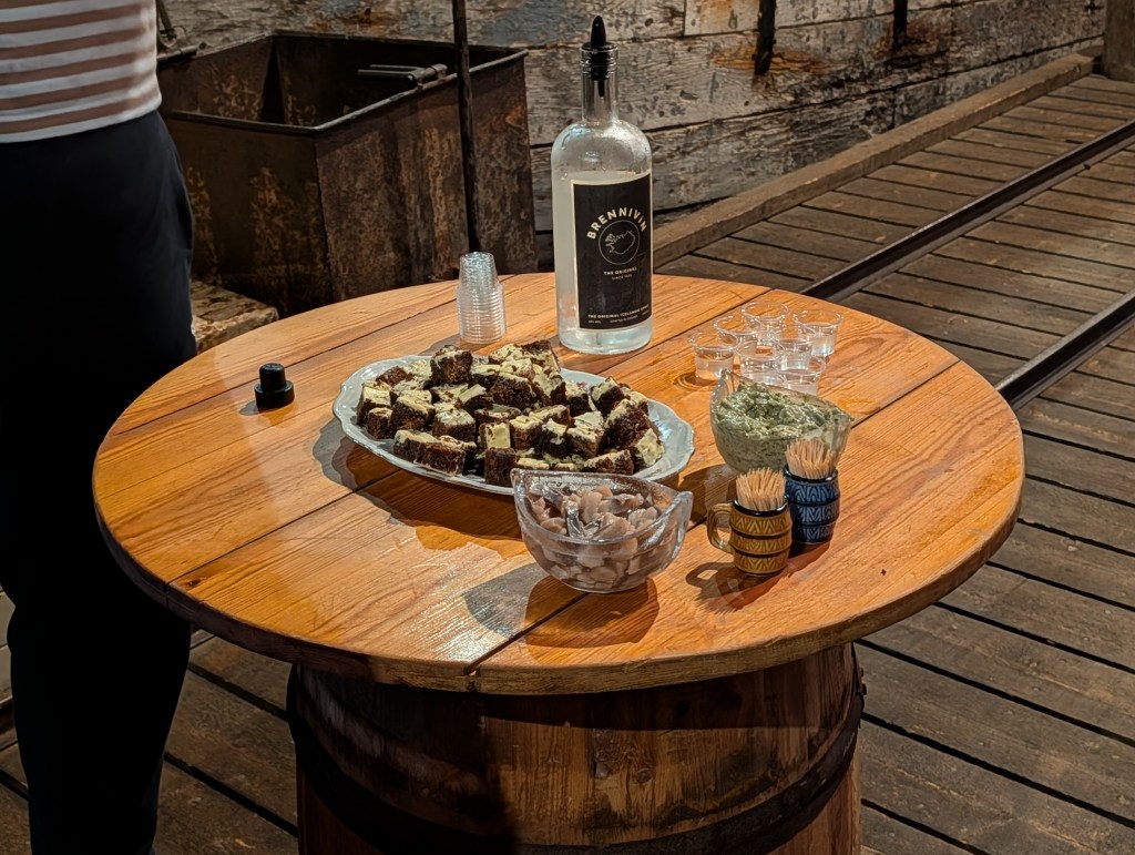 A wooden table displaying a plate of chocolate cake squares, a bottle of Brennivín, shot glasses, a bowl of green dip, and cups with toothpicks.