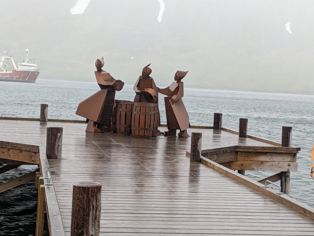 A wooden dock with three bronze sculptures of women interacting around a barrel, and a fishing boat in the background on misty waters.