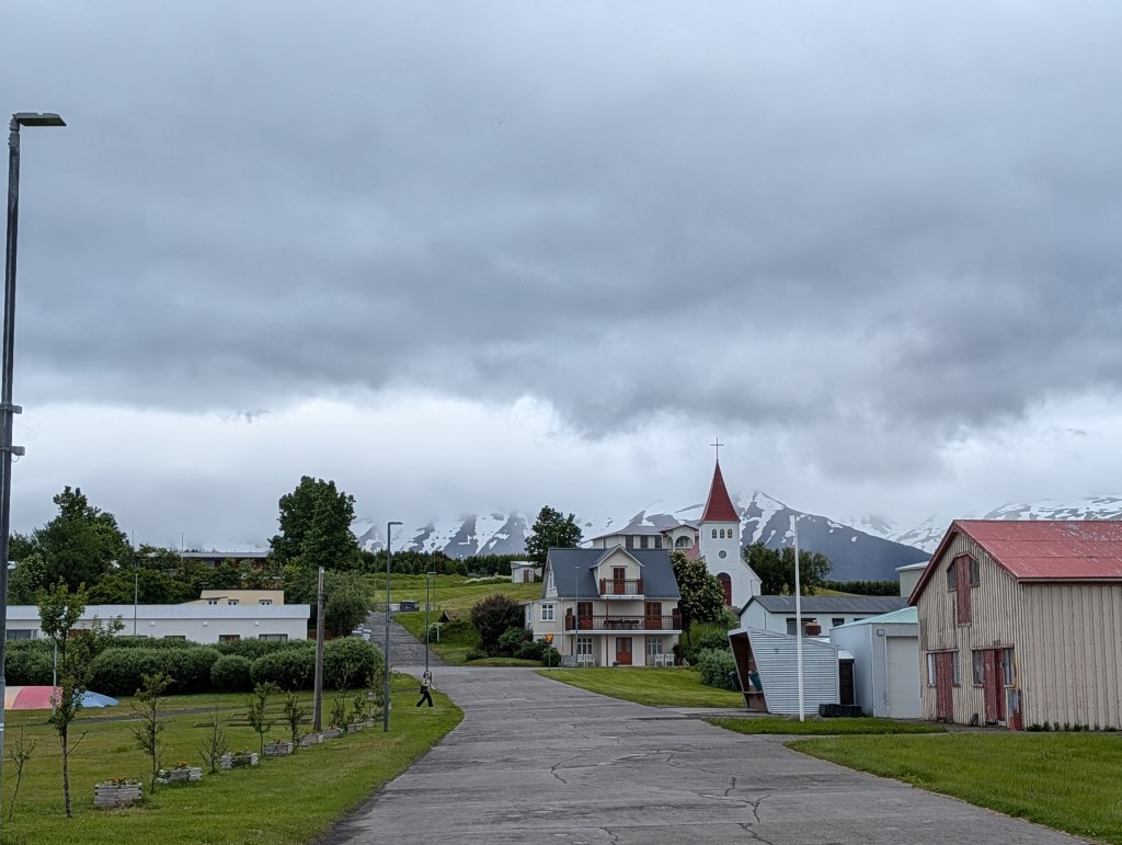A scenic view of a small town featuring a winding road, a white church with a red steeple, colorful houses, and snow-capped mountains in the background under a cloudy sky.