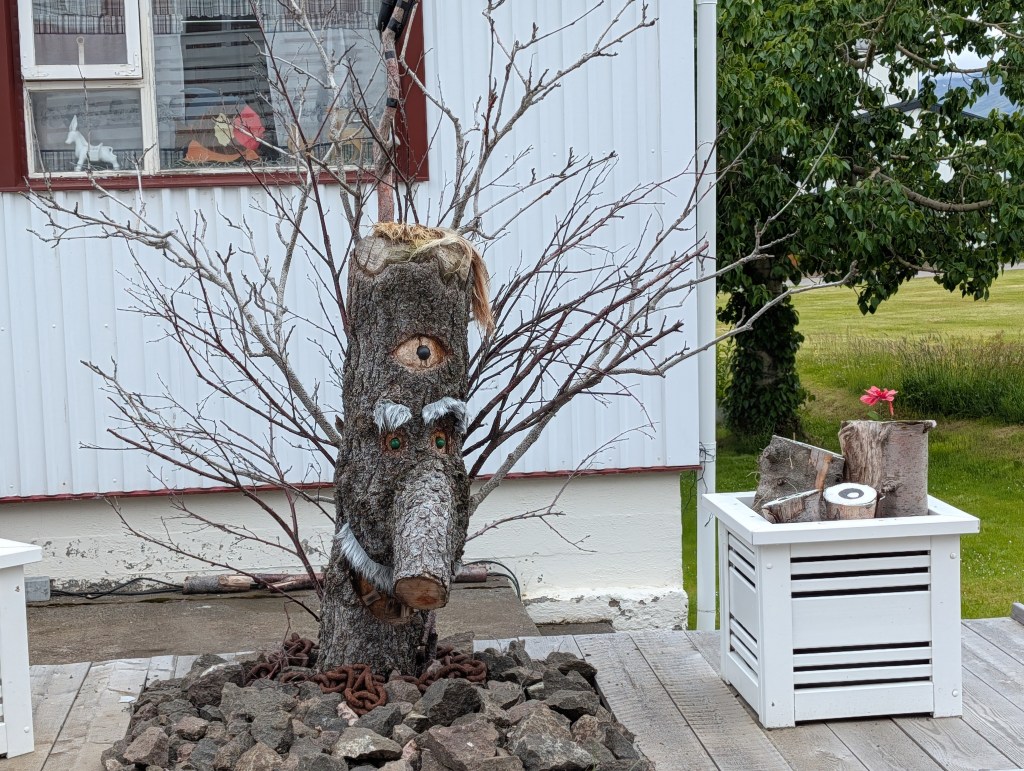 A decorative tree stump sculpture with a carved face, complete with wooden eyes and facial hair, set against a white building and surrounded by stones and planter boxes.