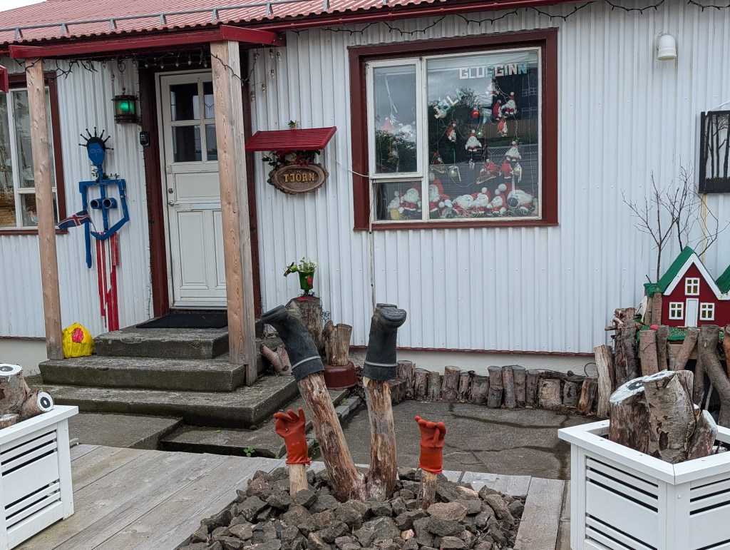 A whimsical outdoor scene featuring a colorful, decorated house with a white facade and red trim. The entrance includes steps leading to a white door, adorned with a blue and red sculpture. The front yard has quirky decorations, including two standing rubber boots and a red glove, surrounded by rocks and wooden logs.