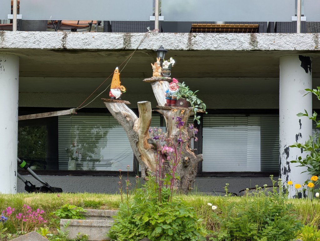 A wooden tree stump decorated with garden gnomes, surrounded by colorful flowers in a residential yard beneath a balcony.