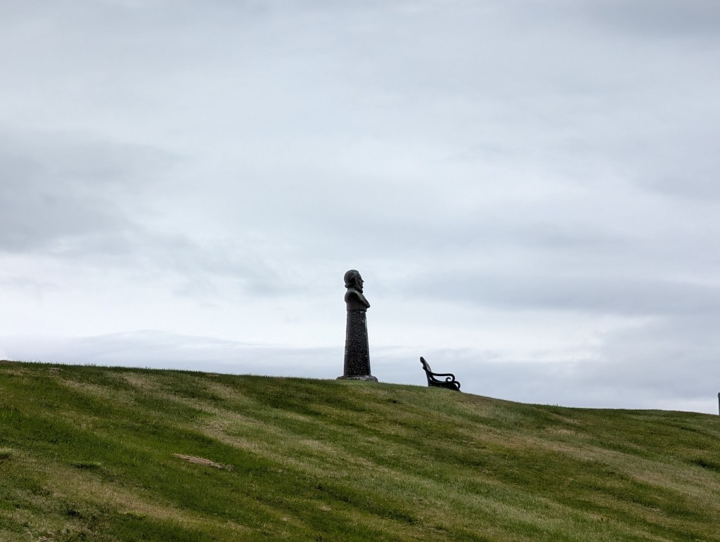 A silhouette of a statue on a grassy hill with a cloudy sky in the background, featuring a bench in the foreground.