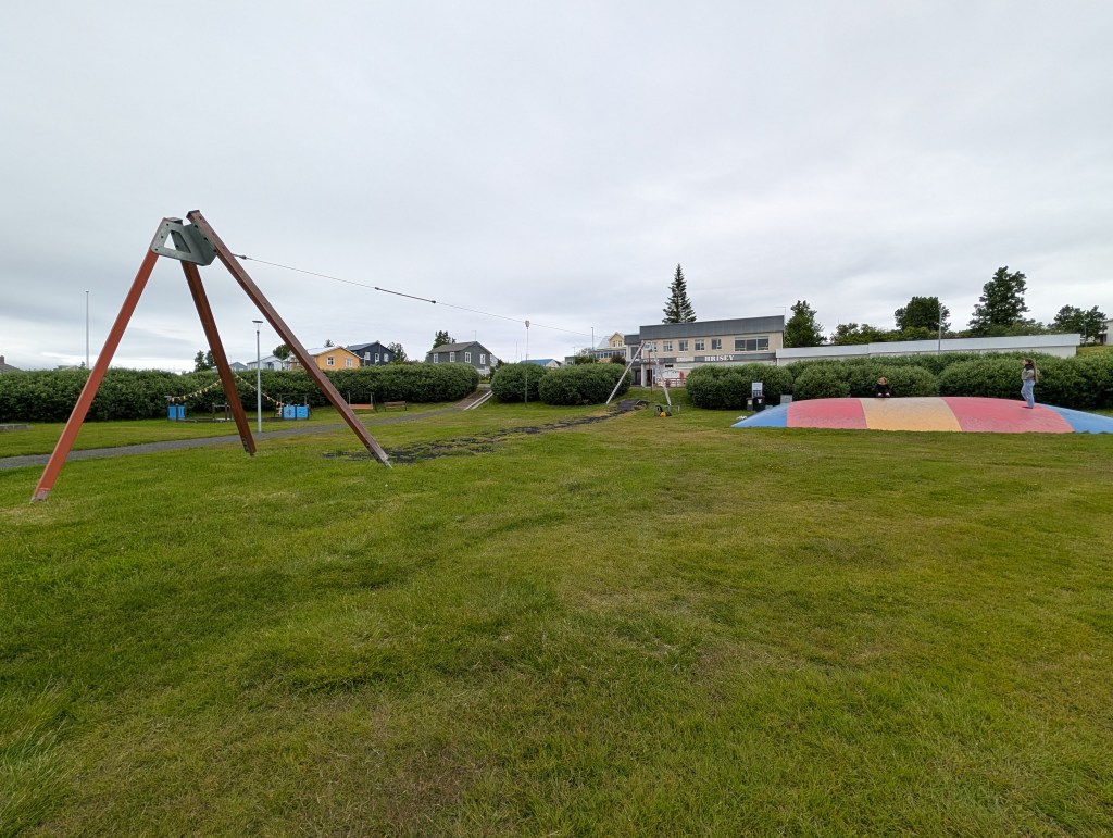 A playground scene featuring a swing set and a colorful jumping pillow, with grassy areas, a building in the background, and cloudy skies.