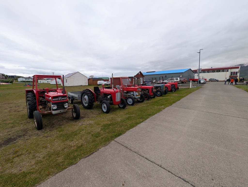 A row of red tractors parked on a grassy area near a building, with overcast skies in the background.