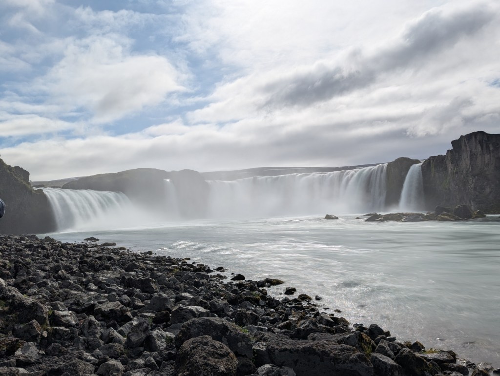 A scenic view of a waterfall cascading over rocky cliffs under a cloudy sky, with a river foreground and smooth water surface.