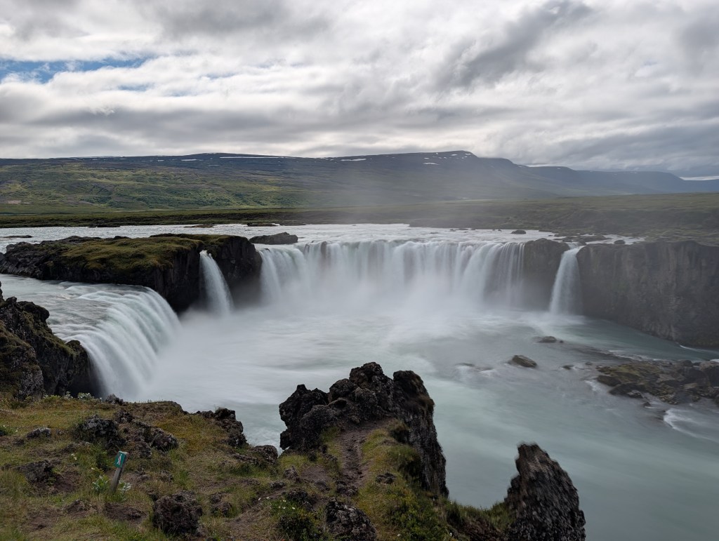 A scenic view of a waterfall cascading over rocky cliffs surrounded by lush greenery and dramatic clouds in the sky.