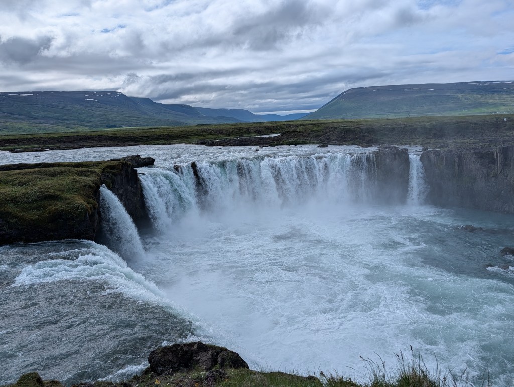 A scenic view of Godafoss waterfall in Iceland, showcasing cascading water over rocky cliffs, surrounded by green landscape and mountains under a cloudy sky.