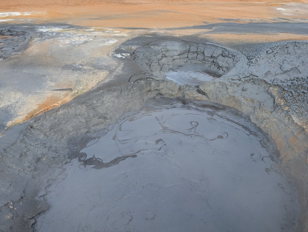 A large mud pot in a geothermal area, showcasing bubbling gray mud with swirling patterns, surrounded by cracked earth and various textures.