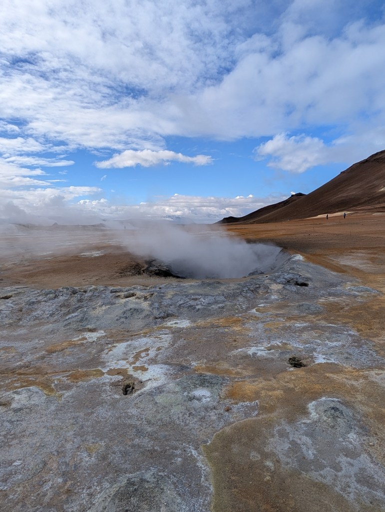 A geothermal area with steam rising from a vent, surrounded by barren land and distant mountains under a partly cloudy sky.